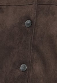 Bruine suède stof met zichtbare textuur; heeft een zwarte viergaatjesknop voor sluiting nabij de naad. Eenvoudig, plat ontwerp.