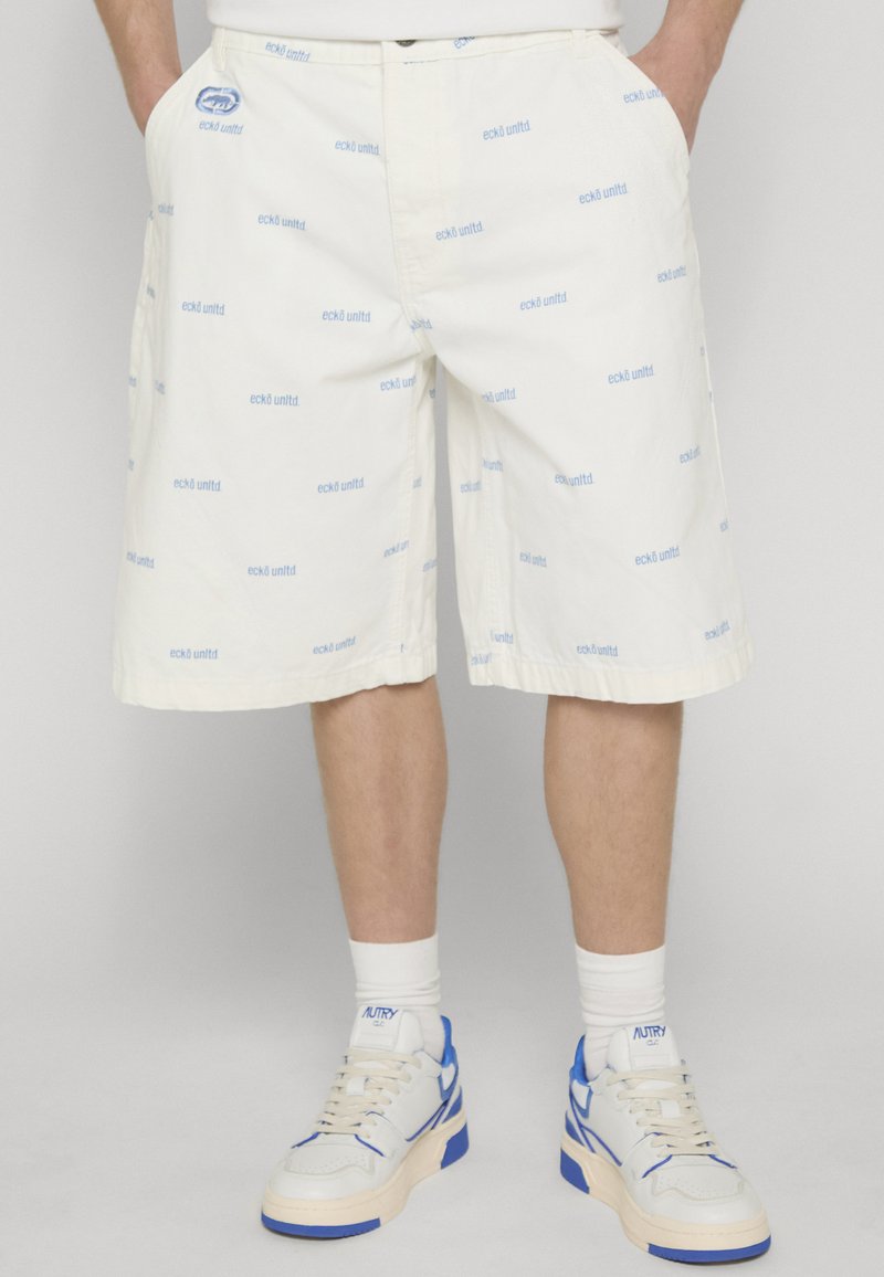 Short blanc avec logos « ecko unltd » bleu clair porté par une personne avec les mains dans les poches, associé à des baskets blanches et bleues et des chaussettes blanches.