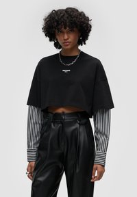 T-shirt cropped nera con logo bianco, sovrapposta a maniche lunghe a righe grigie e bianche. Pantaloni neri in ecopelle a vita alta con pieghe.