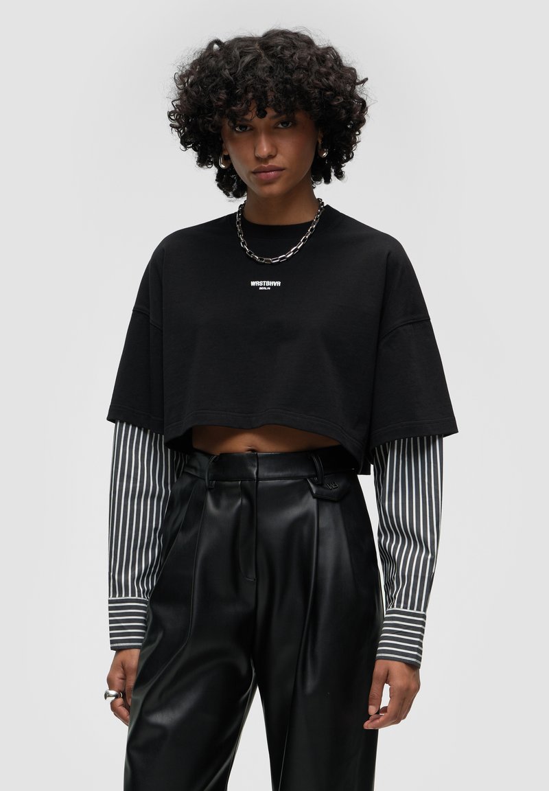 T-shirt cropped nera con logo bianco, sovrapposta a maniche lunghe a righe grigie e bianche. Pantaloni neri in ecopelle a vita alta con pieghe.