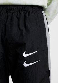 Pantalons de sport noirs avec une taille élastique, ornés de logos Nike blancs, en tissu texturé et avec des panneaux latéraux gris.
