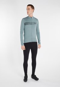 Maillot de cyclisme à manches longues en teal avec une fermeture éclair à l'avant, présentant un design rayé sur la poitrine, associé à des leggings noirs et des chaussures de vélo noires.