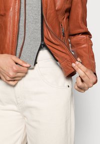 Braune Lederjacke mit grauem Innenreißverschluss und cremefarbener Hose; Detailaufnahme von Händen, die den Reißverschluss justieren. Glatte Textur und figurschmeichelndes Design.