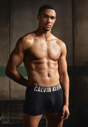 Zwarte boxershorts met een witte "CALVIN KLEIN" logo op de tailleband, gemaakt van een gladde, rekbare stof, gepresenteerd op een gespierde mannelijke model.