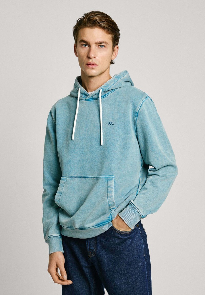 Lichtblauwe hoodie van zachte stof met een voorzak, witte trekkoorden en een klein donker logo op de borst. Gecombineerd met donkere spijkerbroeken.