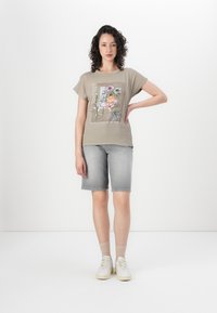 T-shirt à manches courtes khaki avec un imprimé graphique floral et du texte, associé à un short en denim gris. Modèle portant des baskets blanches et des chaussettes claires.