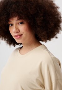 Beige sweatshirt met een ronde halslijn en een ontspannen pasvorm, met een gladde textuur en raglanmouwen, gedragen door een persoon met krullend haar.