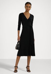 Black wrap dress with three-quarter sleeves, smooth texture, mid-length, and a tied waist detail. Paired with a small black handbag and sandals.