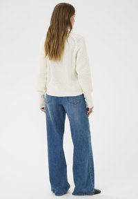 Femme aux longs cheveux bruns portant un pull blanc texturé et un jean bleu à jambes larges, se tenant de dos face à la caméra sur un fond uni.