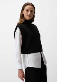Zwarte gebreide vest met een hoge hals, gedragen over een witte overhemd met knopen. Het vest heeft een cropped lengte en losse armopeningen.
