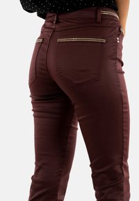 Pantalones burdeos de corte slim, con bolsillos traseros, detalles de cremallera en tono dorado y una textura suave. Tejido elástico para mayor comodidad.