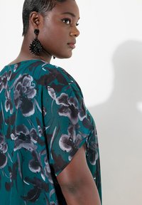 Løs teal top med sort og grå blomstermønster, der har korte ærmer og en løs pasform. Sorte spidse øreringe fuldender looket.