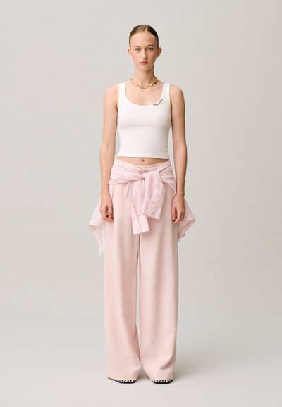 Mujer de pie mirando hacia adelante, lleva una camiseta blanca corta, pantalones anchos de color rosa claro, camisa rosa anudada a la cintura y un collar dorado.