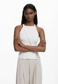 White halter top with a ribbed texture, featuring a diagonal button closure on one side. Paired with light, pleated trousers.