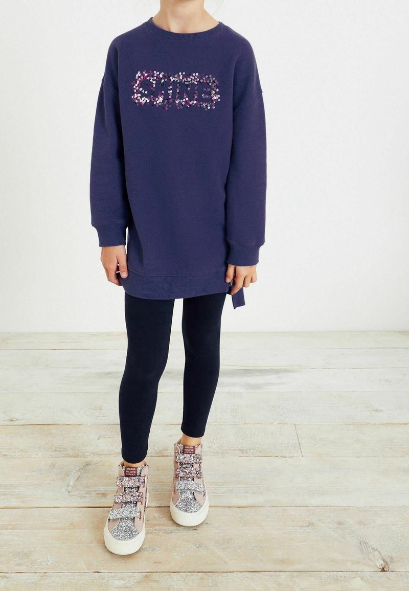 Fialová mikina s nápisem "SHINE" pokrytým barevnými flitry, v kombinaci s černými legínami a světle růžovými třpytivými teniskami. Výrazné textury a akcenty.