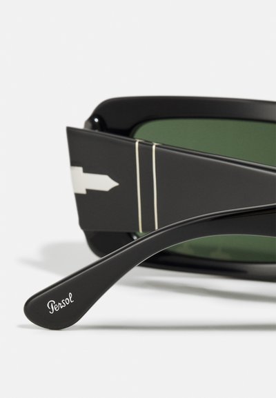 Occhiali da sole con montatura in acetato nero, lenti verdi e un distintivo design a freccia sul lato. Il marchio è visibile sull'asta.