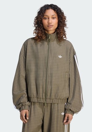 Donna con i capelli ricci che indossa una giacca verde a quadri con cerniera e pantaloni abbinati con strisce bianche sulle maniche e sulle gambe.