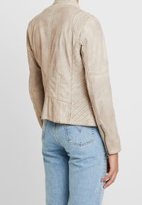 Světle béžová kožená bunda s prošívanými rameny, přiléhavým střihem a zipovými manžetami, v kombinaci s modrými džíny.