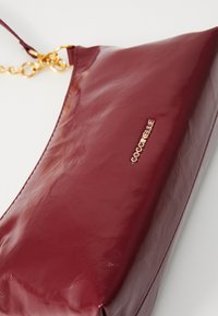 Burgundivörös műbőr válltáska sima textúrával, arany lánc részlettel és domború logóval az elején. Téglalap alakú, minimalista design.