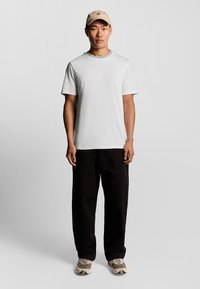 T-shirt bianco a maniche corte, pantaloni neri larghi, cappellino beige con logo e sneakers beige con accenti grigi. Outfit semplice e informale.