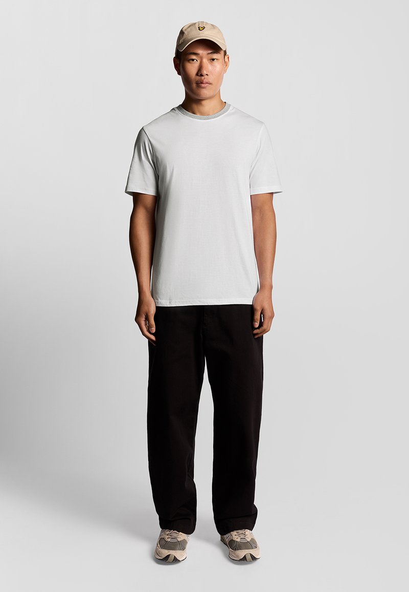 T-shirt bianco a maniche corte, pantaloni neri larghi, cappellino beige con logo e sneakers beige con accenti grigi. Outfit semplice e informale.