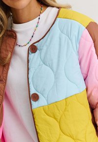 Žena oblečená v mnohofarebnej prešívanej bunde s žltými, modrými, ružovými a hnedými časťami a hnedými gombíkmi, pod ktorou má na sebe biely top a mnohofarebný perlový náhrdelník.