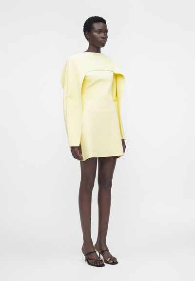 Πρόσωπο με σκουρόχρωμο δέρμα φοράει ένα δομημένο μίνι φόρεμα σε ανοιχτό κίτρινο χρώμα με μακριά, γωνιώδη μανίκια και μαύρα πέδιλα με ανοιχτά δάχτυλα, στέκεται μπροστά σε λευκό φόντο.