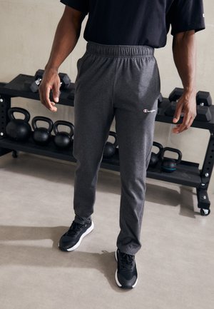 Person trägt dunkelgraue Champion-Sweathose und schwarze Sneakers und steht neben einem Trainingsregal mit Kettlebells und Kurzhanteln.