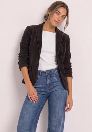Femme portant un blazer en velours côtelé noir par-dessus une chemise blanche et un jean bleu, debout devant un fond clair uni.