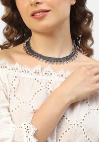 Sort metal choker med lagdelt design, der har buede accenter og hængende dråber. Båret over en hvid broderet off-shoulder top.