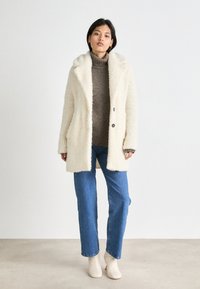 Cream-colored, fluffy coat with a wide collar, two front pockets, and two buttons. Worn over a brown turtleneck and blue jeans.