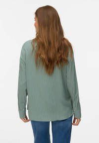 Chemise à manches longues rayée verte à coupe ample, avec un ourlet arrondi et des détails de boutons sur les poignets, associée à un jean bleu.