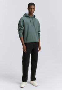 Zöld pamut kapucnis pulóver elülső zsebbel, állítható húzózsinórokkal és bordázott mandzsettával, fekete farmerral és fehér sportcipővel párosítva.