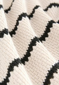 Material tricotat în bej deschis, cu un model în zigzag negru ondulat. Textura este moale, cu cusături vizibile, oferindu-i un aspect modelat și dimensional.