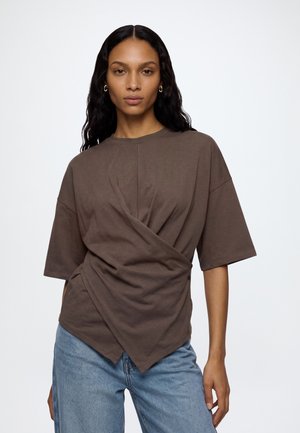 Femme aux longs cheveux noirs portant un haut marron ample à manches courtes style cache-cœur et un jean bleu, debout devant un fond blanc.
