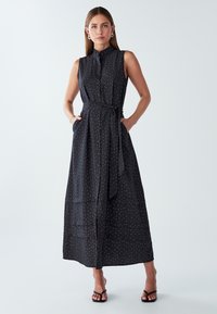 Długa, bezrękawna czarna sukienka w białe kropki, z guzikiem z przodu, paskiem w talii, kieszeniami i warstwowym dołem. Gładka faktura materiału.
