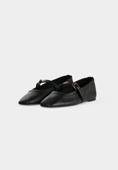 Paire de ballerines noires à bout pointu avec une lanière et une boucle dorée sur chaque chaussure, posée sur un fond clair uni.