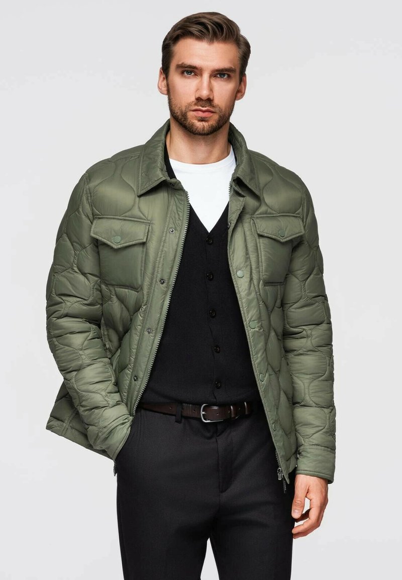 Uomo che indossa un giubbotto trapuntato verde oliva sopra un cardigan nero con bottoni e una maglietta bianca, con pantaloni neri e una cintura marrone, mani in tasca.