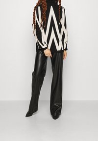 Schwarzer und weißer Pullover mit Zickzackmuster und hohem Kragen, kombiniert mit hochtaillierten schwarzen Lederhosen und spitzen schwarzen Schuhen.