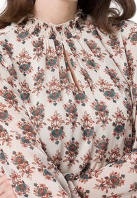 Femme portant une blouse blanche avec des motifs floraux turquoise et marron, un col montant à volants et une découpe en trou de serrure à l'avant.