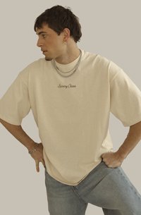 Camiseta de gran tamaño en color beige con bordado de "Luxury Chaos". Presenta mangas cortas y un corte relajado. Combinada con collares de cadena y jeans azul claro.