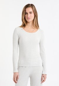 Młoda kobieta w jasnoszarym, prążkowanym długim rękawie i dopasowanych spodniach, stojąca na tle białym.