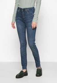 Jeans ajustados de color azul oscuro con una textura suave, con una cinturilla estándar y costuras sutiles, combinados con mocasines verdes.
