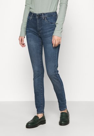 Jeans ajustados de color azul oscuro con una textura suave, con una cinturilla estándar y costuras sutiles, combinados con mocasines verdes.