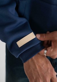 Primer plano de una mano ajustando el puño de un sudadera azul marino con una etiqueta beige que dice "PROHIBIDO".