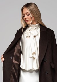 Ruskea villatakki, jossa on kaksi mustaa nappia, esittelee valkoista satiinipaitaa, jossa on iso nauha kaulassa. Sujuva rakenne, mittatilaustyönä valmistettu muotoilu.