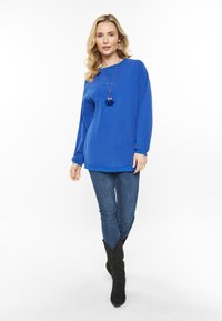 Királykék, hosszúujjú felső kerek nyakkivágással, farmerekkel és fekete térd fölötti csizmával kombinálva. Figyelemre méltó bojt nyaklánc részlet.