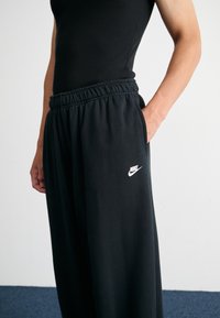 Pantalons de survêtement noirs avec taille élastique et poches latérales, arborant un logo Nike blanc sur le côté gauche. Fabriqués en tissu doux.