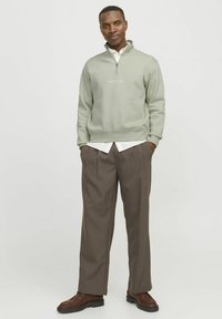 Light green zip-up sweatshirt with white logo, layered over a white collared shirt. Brown pleated trousers and brown boots complete the outfit.