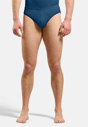 Male legs and lower torso wearing dark blue briefs, standing barefoot against a plain light background.